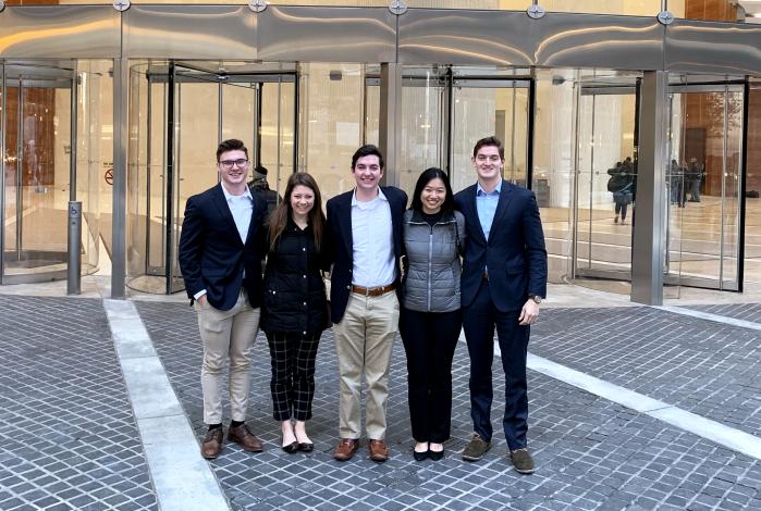 Image: A group of students standing a front of a building's glass entrance. The sign "Deloitte" is visible inside the building.