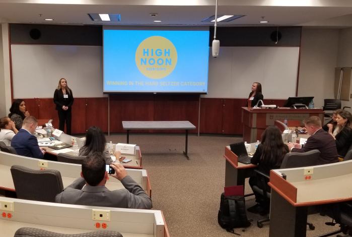 A young woman stands at the front of a lecture-style classroom presenting to those seated around her.